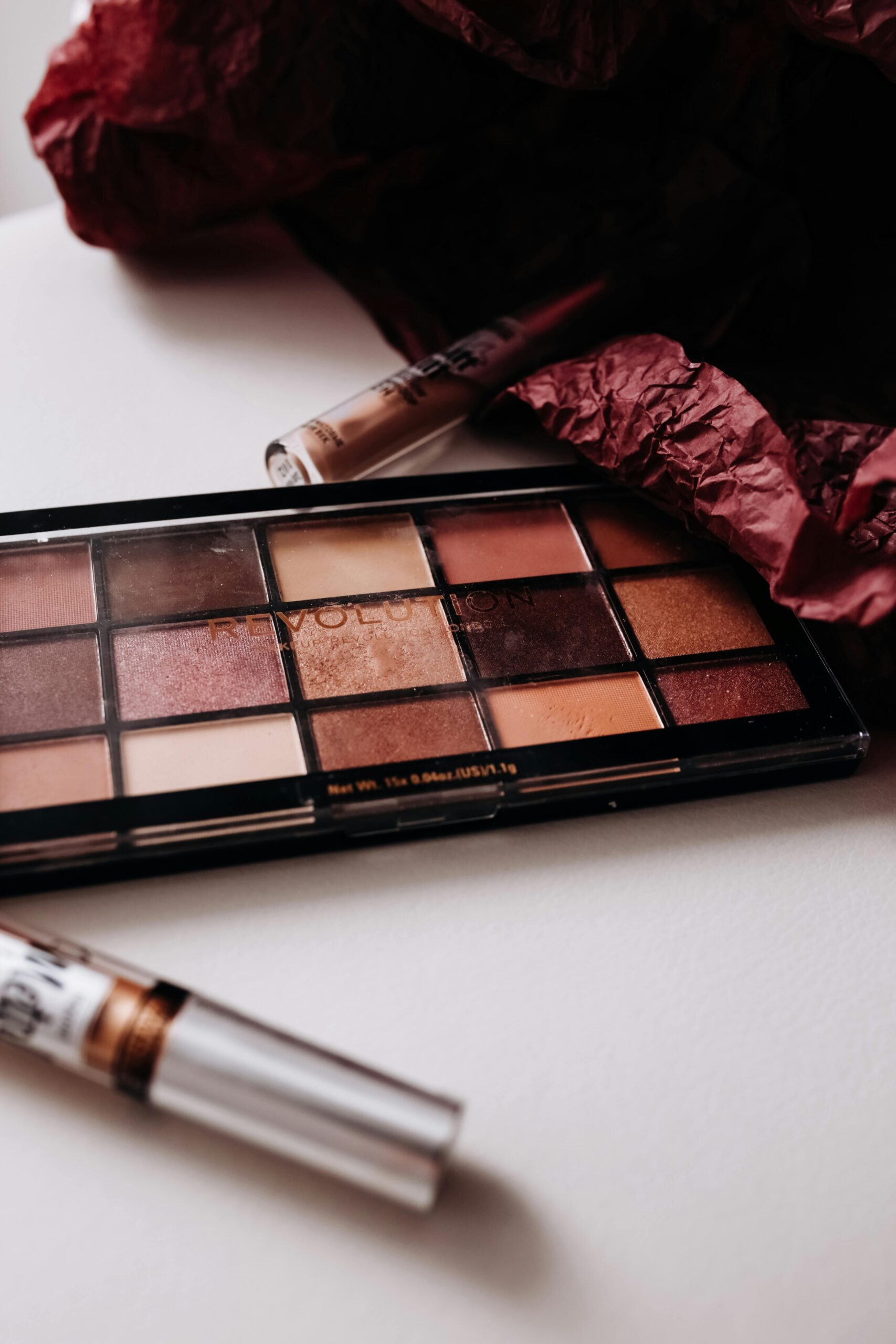 Neutral-toned eyeshadow palette with warm shades, a lip gloss, and makeup products arranged on a white surface with dark red tissue paper.